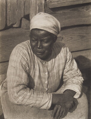 A black-and-white photograph of a Black woman, head covered in a white kerchief, wearing a plaid dress seated on a wood stoop with her arms crossed on her lap.