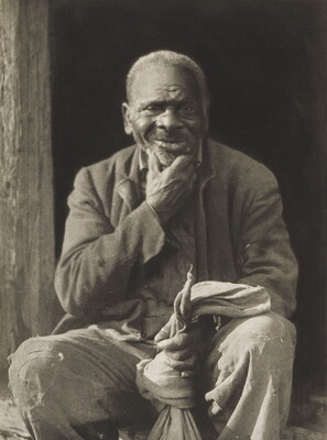 A black-and-white photograph of a seated elderly Black man wearing worn clothing and holding a sack in one hand, the other hand rubbing his stubbled chin.