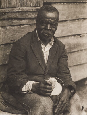 A black-and-white photograph of a seated Black man with a mustache wearing a worn suit jacket holding a lunch pail between his knees.