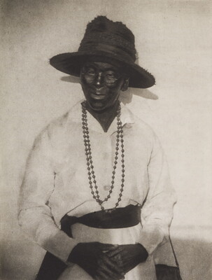 A black-and-white photograph of an older Black woman wearing glasses, a wide-brimmed black hat, and a string of beads over her button-down white blouse.