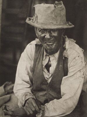 A black-and-white photograph of a seated elderly Black man wearing a tattered hat and a vest over a torn white shirt.