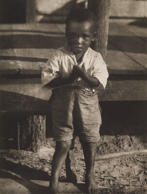 A black-and-white photograph of a young barefoot Black boy wearing a light shirt and dark shorts standing in front of a wood porch.