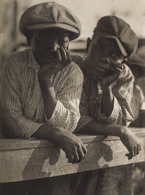 A black-and-white photograph of two young Black boys, both wearing newsboy caps and striped shirts, and both leaning on a wood fence, chins resting in hands.