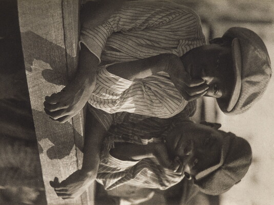A black-and-white photograph of two young Black boys, both wearing newsboy caps and striped shirts, and both leaning on a wood fence, chins resting in hands.