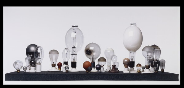 A color photograph of lightbulbs of many shapes and sizes, arranged with their socket sides down on a black platform in front of a white background.