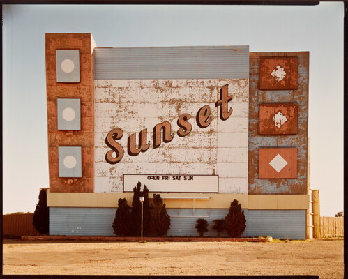A color photograph of a weathered marquee sign for a drive-in movie theater with the word "Sunset" in script.