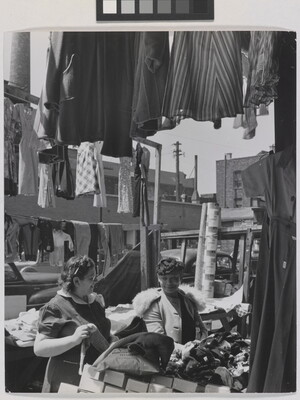 A black-and-white photograph of two light- to medium-skinned women smiling at each other in an open-air market, surrounded by clothing and boxes.