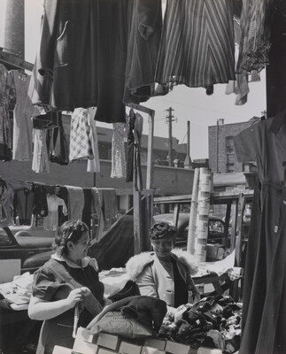 A black-and-white photograph of two light- to medium-skinned women smiling at each other in an open-air market, surrounded by clothing and boxes.
