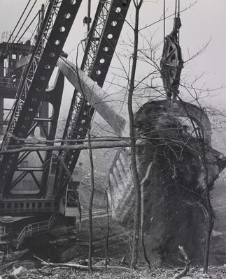 A black-and-white photograph of a massive digging machine taking a huge chunk of earth out of the ground with spindly trees in the foreground.