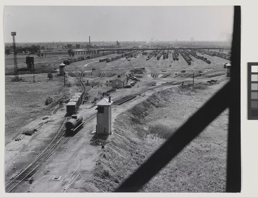 A black-and-white photograph of several train cars on tracks that lead to a train yard filled with more cars in the distance.