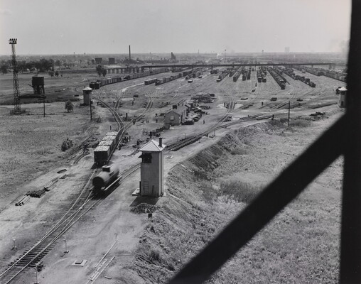 A black-and-white photograph of several train cars on tracks that lead to a train yard filled with more cars in the distance.