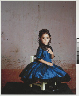 A color photograph of a young girl in a formal blue dress posed on a white chair.