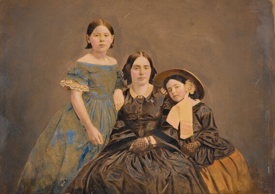 A color photograph of a seated White woman with two girls, one standing on either side of her, all wearing long voluminous dresses.