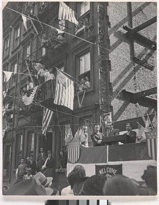 A black-and-white photograph of a man making a speech on a platform with people watching and American flags hanging from apartment windows above.
