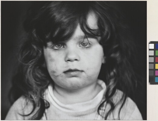 A black-and-white photograph of a White, toddler girl with dark, curly hair and dirt on her cheeks looking directly at the viewer.