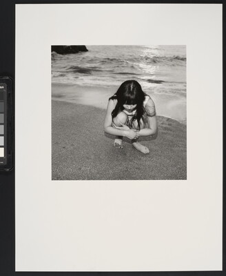 A black-and-white photograph of a young White girl with long dark hair crouched on a shoreline looking at the sand.