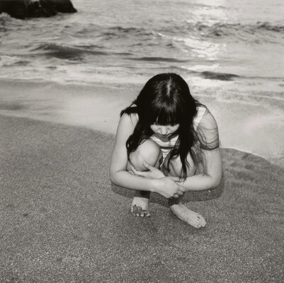 A black-and-white photograph of a young White girl with long dark hair crouched on a shoreline looking at the sand.