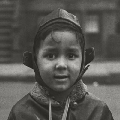 A detail of a black-and-white photograph of a warmly dressed young boy with medium-toned skin wearing a drum from his neck and looking directly into camera.