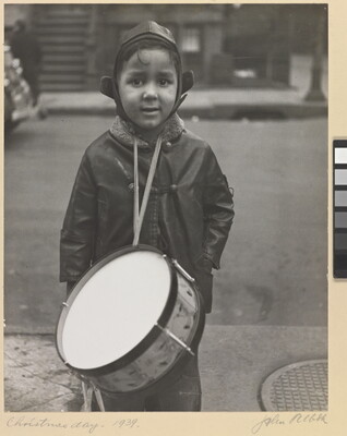 A black-and-white photograph of a warmly dressed young boy with medium-toned skin wearing a drum from his neck and looking directly into camera.