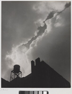 A black-and-white photograph taken from the ground looking up of silhouettes of industrial buildings and a water tower against a cloudy sky.