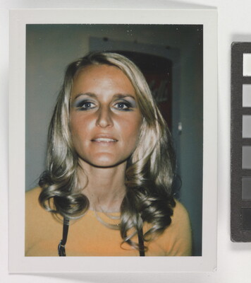An informal color photograph of a White woman from the shoulders up, with blonde hair, blue eyeshadow, and a tanned face.