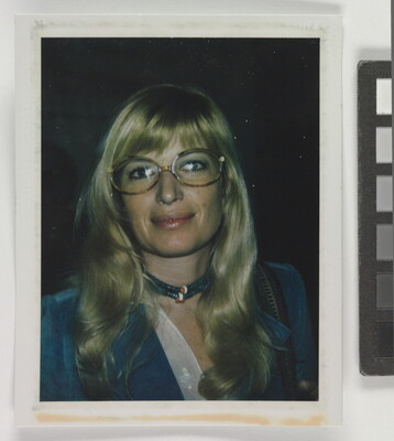 An informal color photograph of a White woman with blonde hair and tortoise-shell glasses wearing a blue suit and necklace.