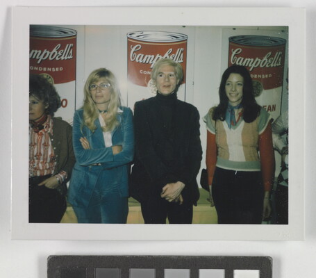 A color photograph of four White people standing in front of three iconic images of "Campbell's Soup" cans.