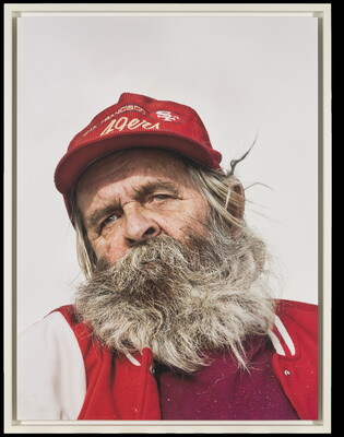 A color photograph of an older White man with a scraggly gray beard wearing a red 49ers hat and a red jacket.