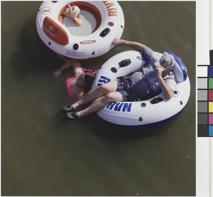 A color photograph taken from above of a White man reclining in an innertube and a White woman in pink swimsuit soaking in the water next to an innertube.