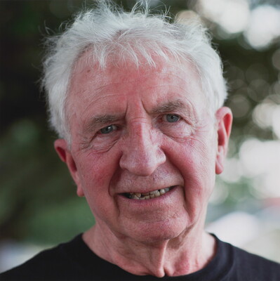 A color photograph of the head and shoulders of a smiling older White man with white tousled hair.