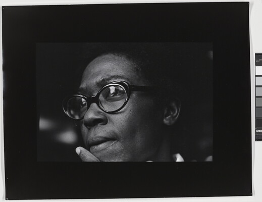 A black-and-white close-up photograph of the face of a Black woman wearing black glasses, an index finger held next to her mouth.