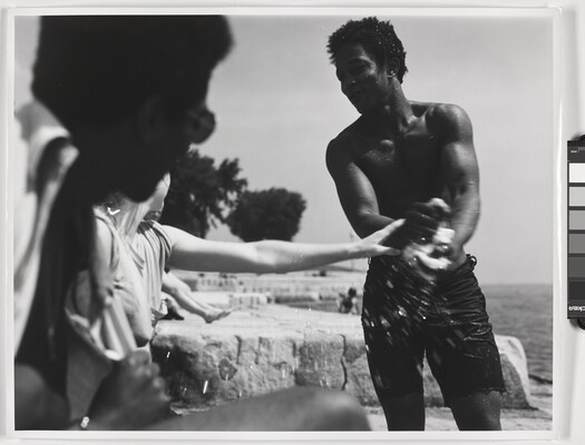 A black-and-white photograph of a smiling Black man in swim shorts wringing water out of something as a White woman reaches toward him.