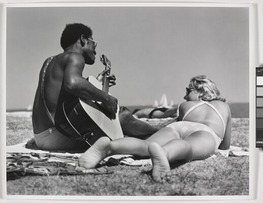 A black-and-white photograph of a Black man playing guitar on a beach blanket next to a White woman lying on her stomach with her feet toward the camera.