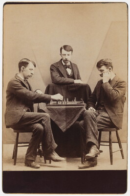 A sepia-toned photograph of the same White man with a moustache wearing a suit seated in three different positions around a chess board.