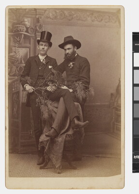 A sepia-toned studio photograph of two White men in suits, each standing with one a leg over the back of a single chair so that their legs overlap.