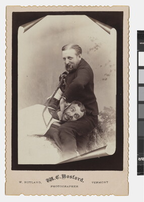 A sepia-toned studio photograph of a seated White man holding a saw against the neck of another White man whose head is resting on his lap.