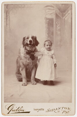 A black-and-white studio photograph of a White toddler in a white dress standing next to a large dog and holding onto its collar. 