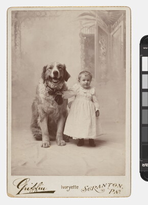 A black-and-white studio photograph of a White toddler in a white dress standing next to a large dog and holding onto its collar. 
