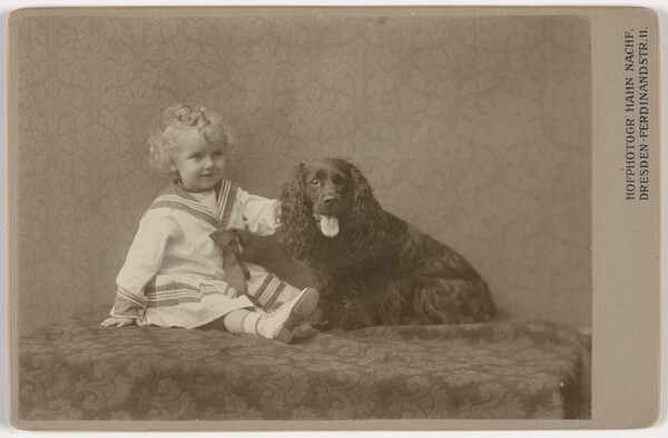 A sepia-toned photograph of a young White child with blond ringlets wearing a sailor dress sitting next to an Irish setter-like dog.