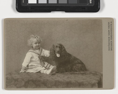 A sepia-toned photograph of a young White child with blond ringlets wearing a sailor dress sitting next to an Irish setter-like dog.