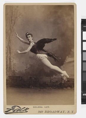A sepia-toned studio photograph of a White woman, black wings on her back, arms extended, suspended in the air as if flying.