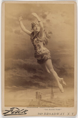A sepia-toned studio photograph of a White woman dressed as a fairy suspended in the air as if flying over a city skyline.