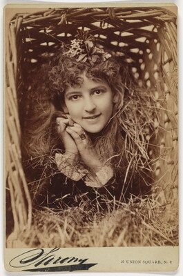 A sepia-toned photograph of the head and shoulders of a smiling White woman that looks as if she is in a wicker basket filled with straw.