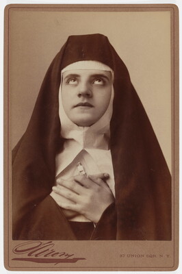 A black-and-white photograph of a White nun wearing a full habit, including a veil, coif, and wimple, holding her hands to her chest and looking up.