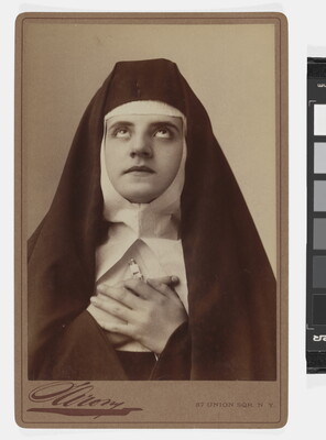 A black-and-white photograph of a White nun wearing a full habit, including a veil, coif, and wimple, holding her hands to her chest and looking up.