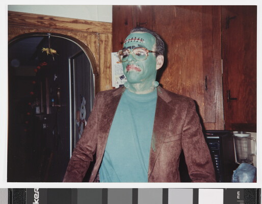 A color photograph of a man wearing green face paint and a Frankenstein costume standing in what looks like a kitchen.