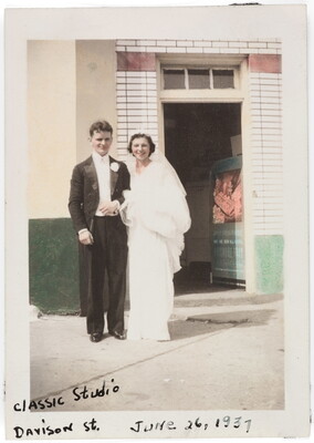 A color photograph of a young White man and woman dressed in wedding attire and standing in front of a doorway.