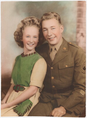 A color photograph of a White woman with curly blonde hair wearing a green and white dress sitting next to a White man in a green military uniform.