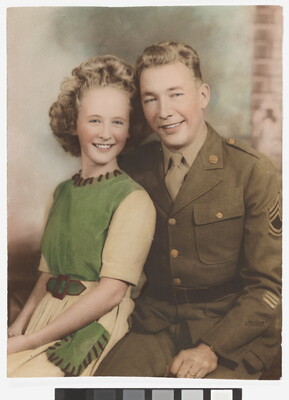 A color photograph of a White woman with curly blonde hair wearing a green and white dress sitting next to a White man in a green military uniform.