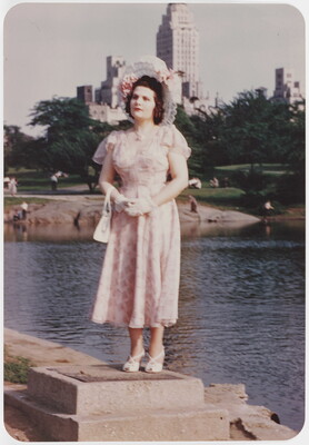 A color photograph of a White woman wearing a decorative sunhat and a floral dress standing on a concrete block beside a pond with tall buildings in the background.